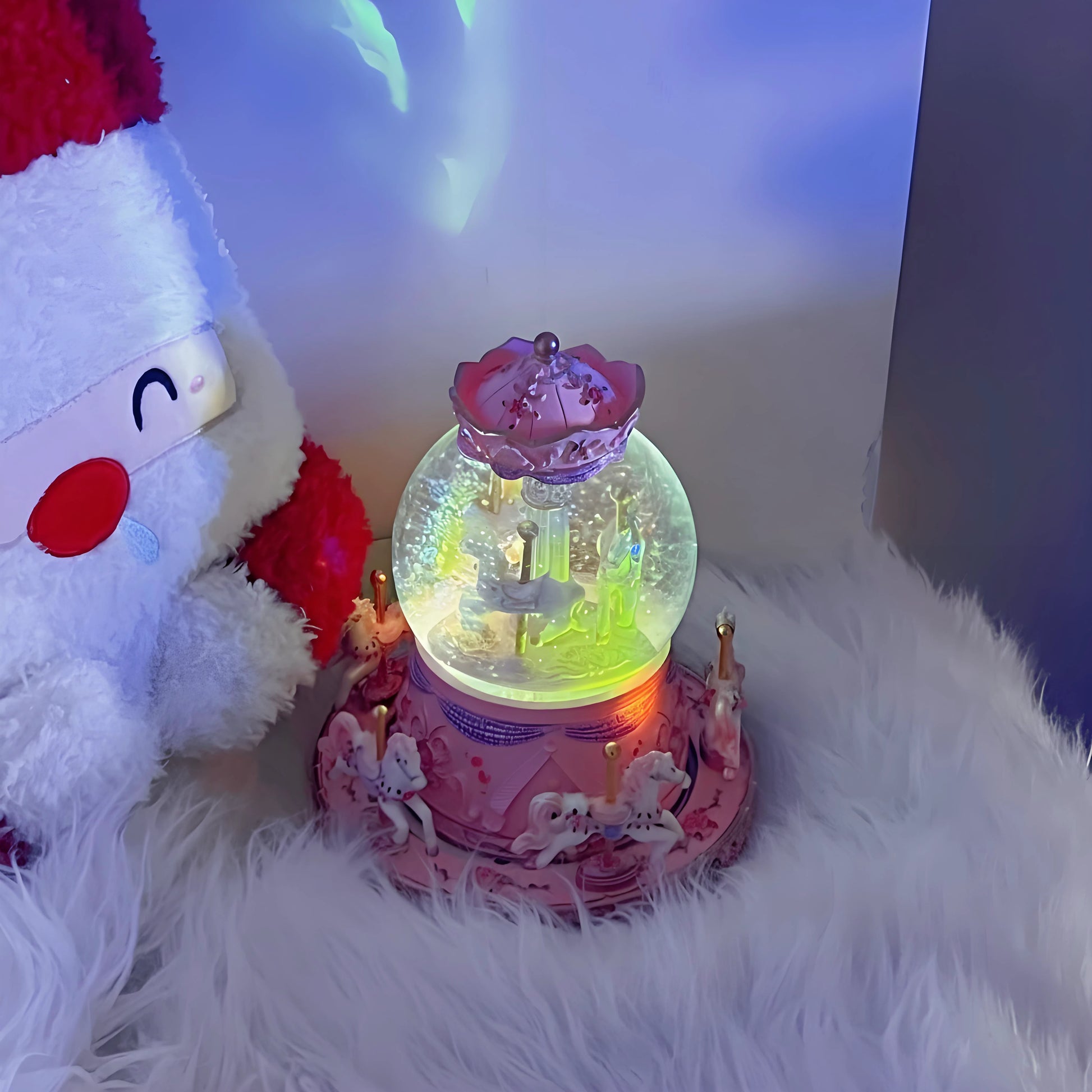 Decorative snow globe with a pink base and fairy on a white fluffy surface next to a plush toy.
