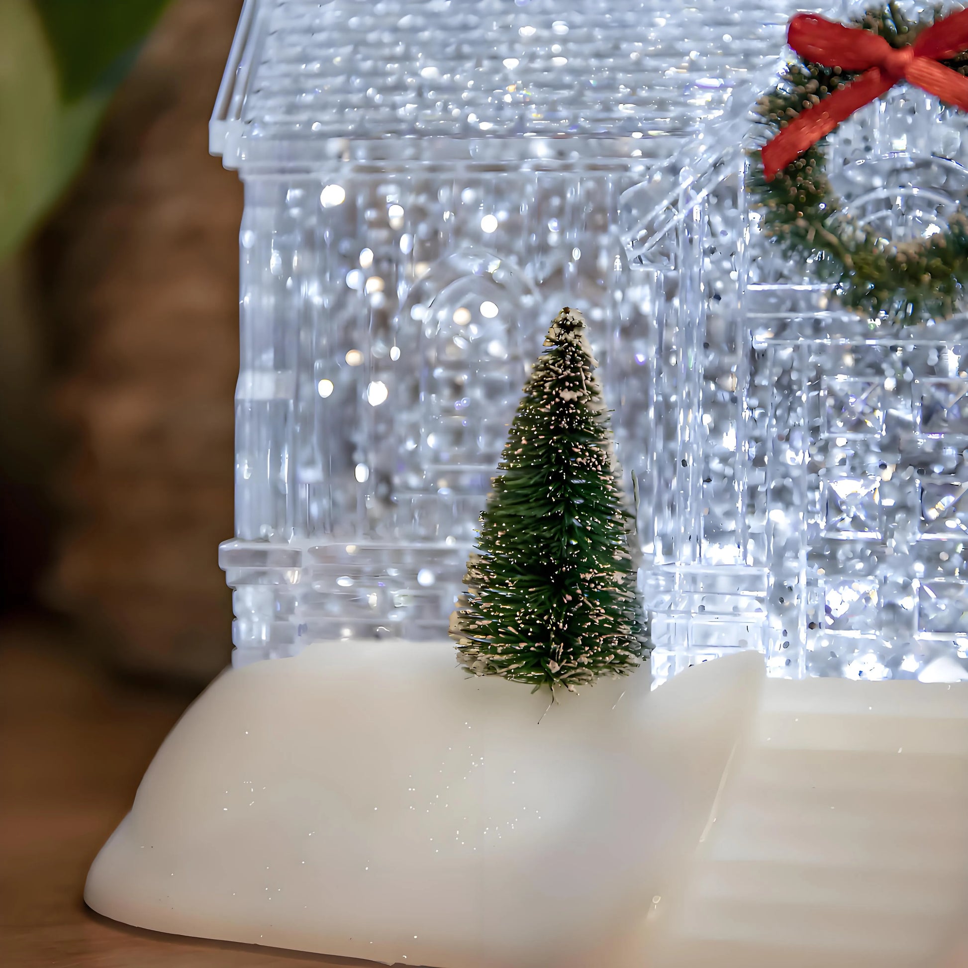 Decorative Christmas scene with a clear crystal house, small tree, and wreath on a wooden surface.